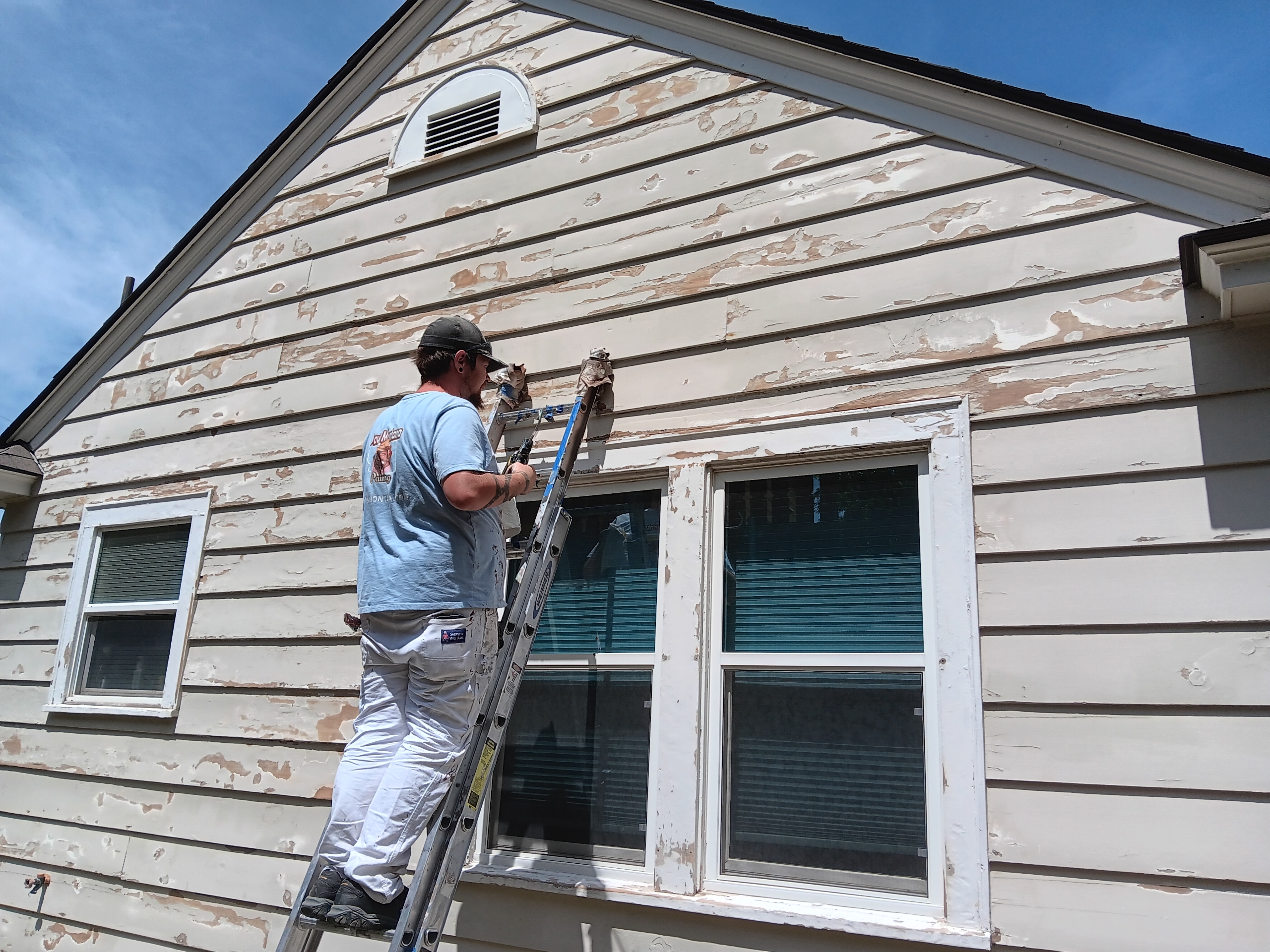 A painter preparing white siding on a ladder